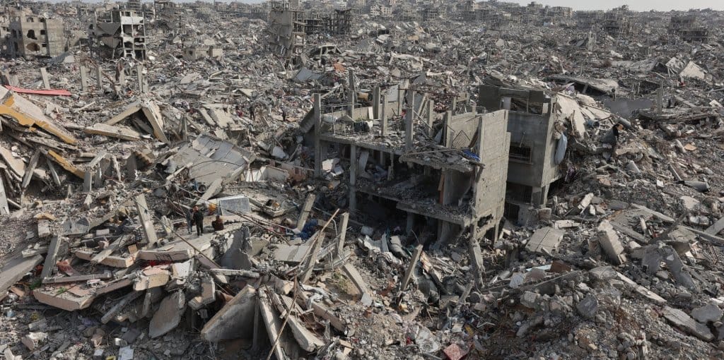 “ La imagen muestra un vasto paisaje de escombros y edificios derrumbados, hundidos en escombros. Se ven estructuras de hormigón rotas y una atmósfera de desconsoladora destrucción. Jabaliya, 19 de enero de 2025. Esta vista aérea muestra a palestinos desplazados revisando las ruinas de una casa a su regreso al campo de refugiados devastado por la guerra de Jabaliya, en el norte de la Franja de Gaza. (Omar AL-QATTAA / AFP) 